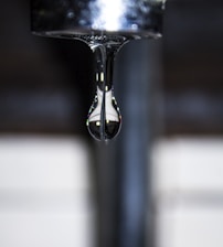 A single water droplet is suspended in mid-air, hanging from a shiny metallic tap. The background is out of focus, emphasizing the clarity and detail of the droplet, which reflects light and surroundings.