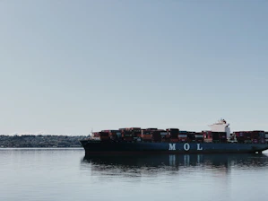 A large cargo ship loaded with colorful containers sailing on calm ocean waters under a clear sky.