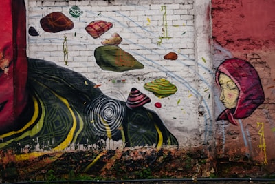 A vibrant mural painted on a weathered brick wall. The artwork includes abstract elements such as floating stones in various shapes and colors. There is a figure of a woman wearing a red hood, portrayed with a calm expression. The mural also features bold, swirling yellow and green patterns near the bottom and a stylized series of concentric circles.