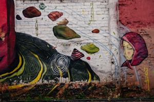 A vibrant mural painted on a weathered brick wall. The artwork includes abstract elements such as floating stones in various shapes and colors. There is a figure of a woman wearing a red hood, portrayed with a calm expression. The mural also features bold, swirling yellow and green patterns near the bottom and a stylized series of concentric circles.