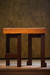 A wooden table with a thick, textured tabletop supported by sturdy legs, set against a blurred, earthy-toned background. The table is positioned on a wooden floor, and the lighting casts soft shadows underneath.