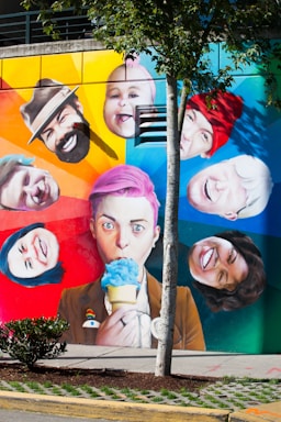 A vibrant and colorful mural depicting various expressive faces surrounding a central figure with pink hair holding a blue ice cream cone. The background features a rainbow gradient, and a tree partially obscures the view. Each face exhibits a different expression, adding a playful and dynamic feel to the scene.