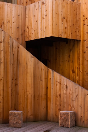 A wooden architectural structure featuring large, vertical planks with a warm, natural finish. Two cubic wooden stools sit on a wooden deck, suggesting a minimalist and rustic design. The geometric forms create an interesting visual composition.