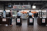 A bustling train station platform with people waiting for a train. Two uniformed officers stand at ticket gates, overseeing the platform. An electronic board displays train times and destinations, and a clock shows the current time. The train in the background is orange with silver details.