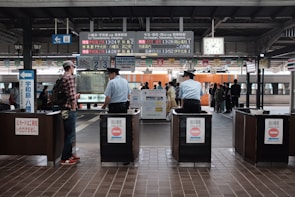 A bustling train station platform with people waiting for a train. Two uniformed officers stand at ticket gates, overseeing the platform. An electronic board displays train times and destinations, and a clock shows the current time. The train in the background is orange with silver details.