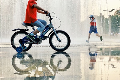 A child is riding a blue bicycle with training wheels near a water fountain, while another child wearing a red, white, and blue outfit runs through the fountain. The ground is wet, creating reflections of the children and the fountain.