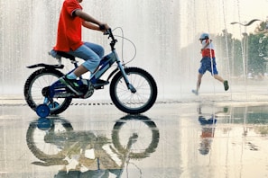 A child is riding a blue bicycle with training wheels near a water fountain, while another child wearing a red, white, and blue outfit runs through the fountain. The ground is wet, creating reflections of the children and the fountain.