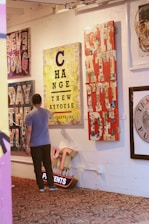 A man observes several vibrant and colorful artworks displayed on a wall in a gallery setting. The artworks feature bold typography and mixed media designs with messages like 'CHANGE' and 'GRATITUDE'. The floor is covered with an ornate patterned carpet, and there is a neon sign on the floor.