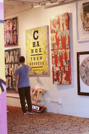A man observes several vibrant and colorful artworks displayed on a wall in a gallery setting. The artworks feature bold typography and mixed media designs with messages like 'CHANGE' and 'GRATITUDE'. The floor is covered with an ornate patterned carpet, and there is a neon sign on the floor.
