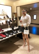 A stylishly dressed individual is standing in a shoe store. They are wearing oversized sunglasses, a sequined blouse, a black skirt with white dots, and clear high-heeled sandals. The person carries a white designer handbag with a gold chain strap and holds a glass of wine. The store features a variety of fashion-forward shoes on shelves, including boots and chunky loafers in assorted colors.