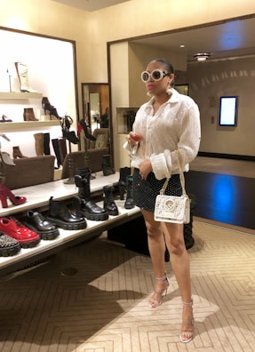 A stylishly dressed individual is standing in a shoe store. They are wearing oversized sunglasses, a sequined blouse, a black skirt with white dots, and clear high-heeled sandals. The person carries a white designer handbag with a gold chain strap and holds a glass of wine. The store features a variety of fashion-forward shoes on shelves, including boots and chunky loafers in assorted colors.