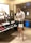 A stylishly dressed individual is standing in a shoe store. They are wearing oversized sunglasses, a sequined blouse, a black skirt with white dots, and clear high-heeled sandals. The person carries a white designer handbag with a gold chain strap and holds a glass of wine. The store features a variety of fashion-forward shoes on shelves, including boots and chunky loafers in assorted colors.