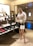 A stylishly dressed individual is standing in a shoe store. They are wearing oversized sunglasses, a sequined blouse, a black skirt with white dots, and clear high-heeled sandals. The person carries a white designer handbag with a gold chain strap and holds a glass of wine. The store features a variety of fashion-forward shoes on shelves, including boots and chunky loafers in assorted colors.