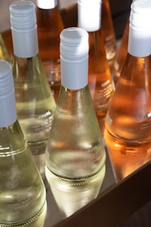 Several bottles of wine are neatly arranged in a wooden tray, showcasing both clear and amber liquids. Each bottle features a white cap and a minimalist design. The sunlight casts a bright reflection on the glass, enhancing the clarity and highlights of the bottles.