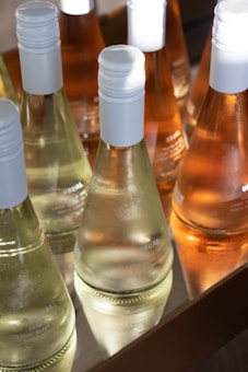 Several bottles of wine are neatly arranged in a wooden tray, showcasing both clear and amber liquids. Each bottle features a white cap and a minimalist design. The sunlight casts a bright reflection on the glass, enhancing the clarity and highlights of the bottles.