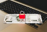 An Indian 500 rupee note is placed on a laptop keyboard with a red padlock sitting on top of the note.