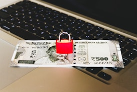 An Indian 500 rupee note is placed on a laptop keyboard with a red padlock sitting on top of the note.