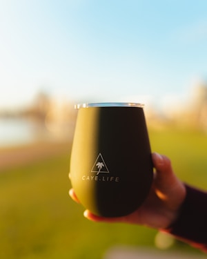 A hand holds a matte black tumbler with the logo 'Caye.Life' featuring a palm tree inside a triangle. In the background, there is an out-of-focus outdoor scene with green grass and a bright blue sky.