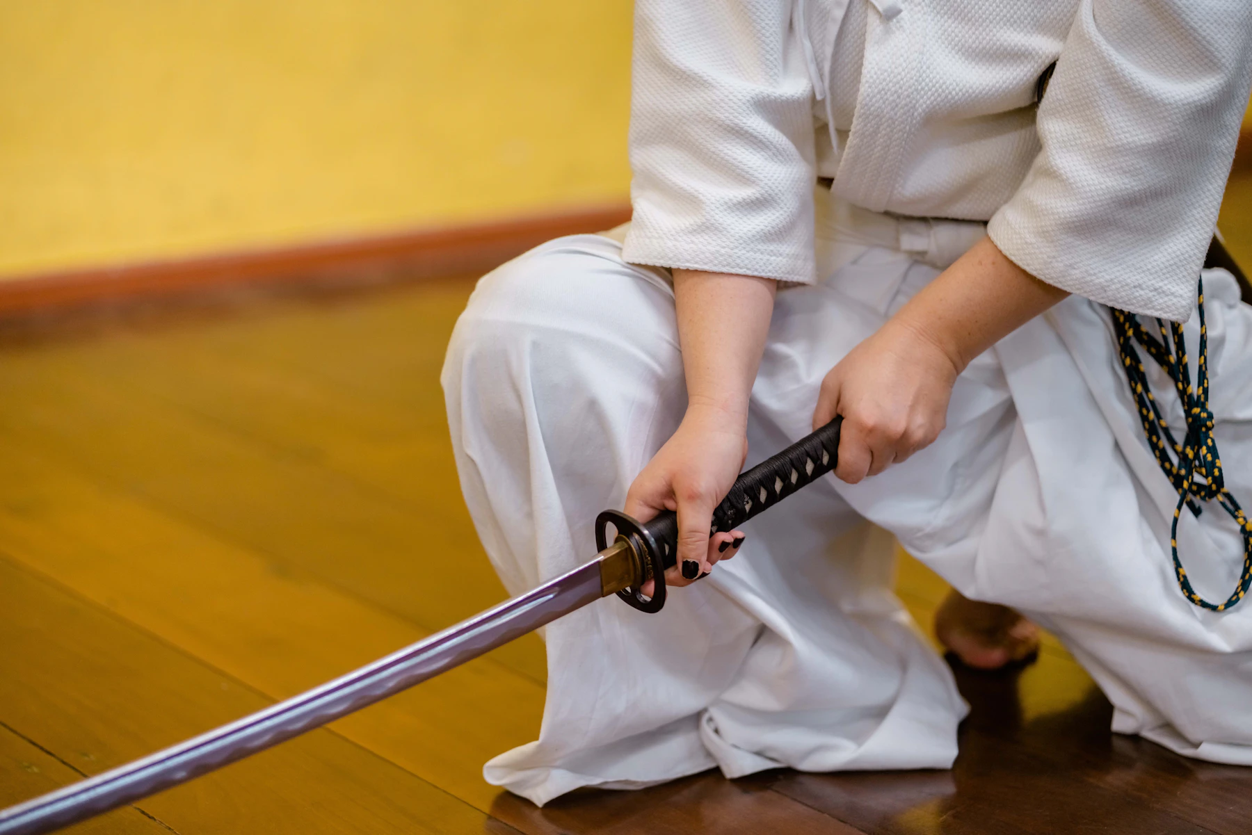 A kneeling person in white holding a drawn katana by the grip above a wooden floor