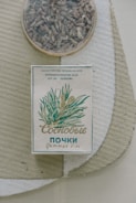 An old-style package of herbal tea or medicine is placed on a textured surface. The box features plant illustrations and Cyrillic text, likely indicating its origin and purpose. A round container with dried herbs or loose tea sits nearby.