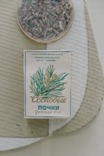 An old-style package of herbal tea or medicine is placed on a textured surface. The box features plant illustrations and Cyrillic text, likely indicating its origin and purpose. A round container with dried herbs or loose tea sits nearby.