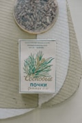 An old-style package of herbal tea or medicine is placed on a textured surface. The box features plant illustrations and Cyrillic text, likely indicating its origin and purpose. A round container with dried herbs or loose tea sits nearby.