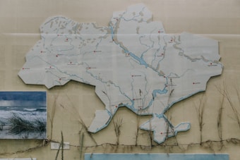 An illustrative map of Ukraine features various rivers and cities marked with red circles. A photographic image of coastal grass and waves is placed to the left. The map is presented on a neutral background, with dried grass stems placed underneath.