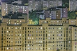 A cluster of residential apartment buildings at night with numerous illuminated windows. The structures are uniform and consist of multiple stories, situated against a backdrop of dark trees and dimly lit streets.
