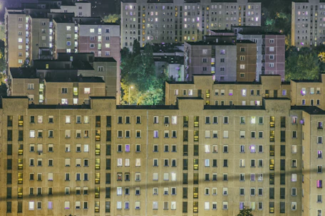 A cluster of residential apartment buildings at night with numerous illuminated windows. The structures are uniform and consist of multiple stories, situated against a backdrop of dark trees and dimly lit streets.