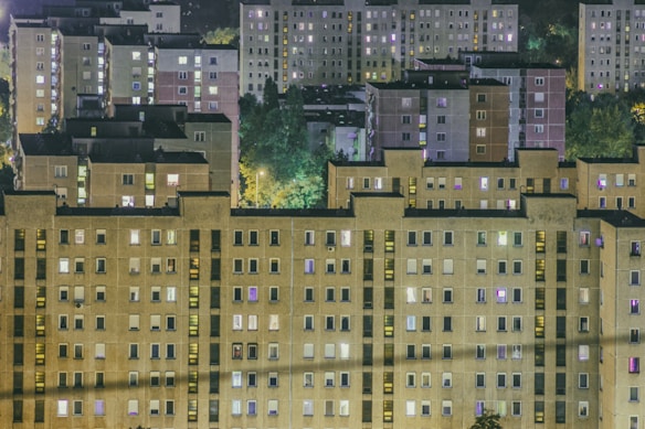 A cluster of residential apartment buildings at night with numerous illuminated windows. The structures are uniform and consist of multiple stories, situated against a backdrop of dark trees and dimly lit streets.