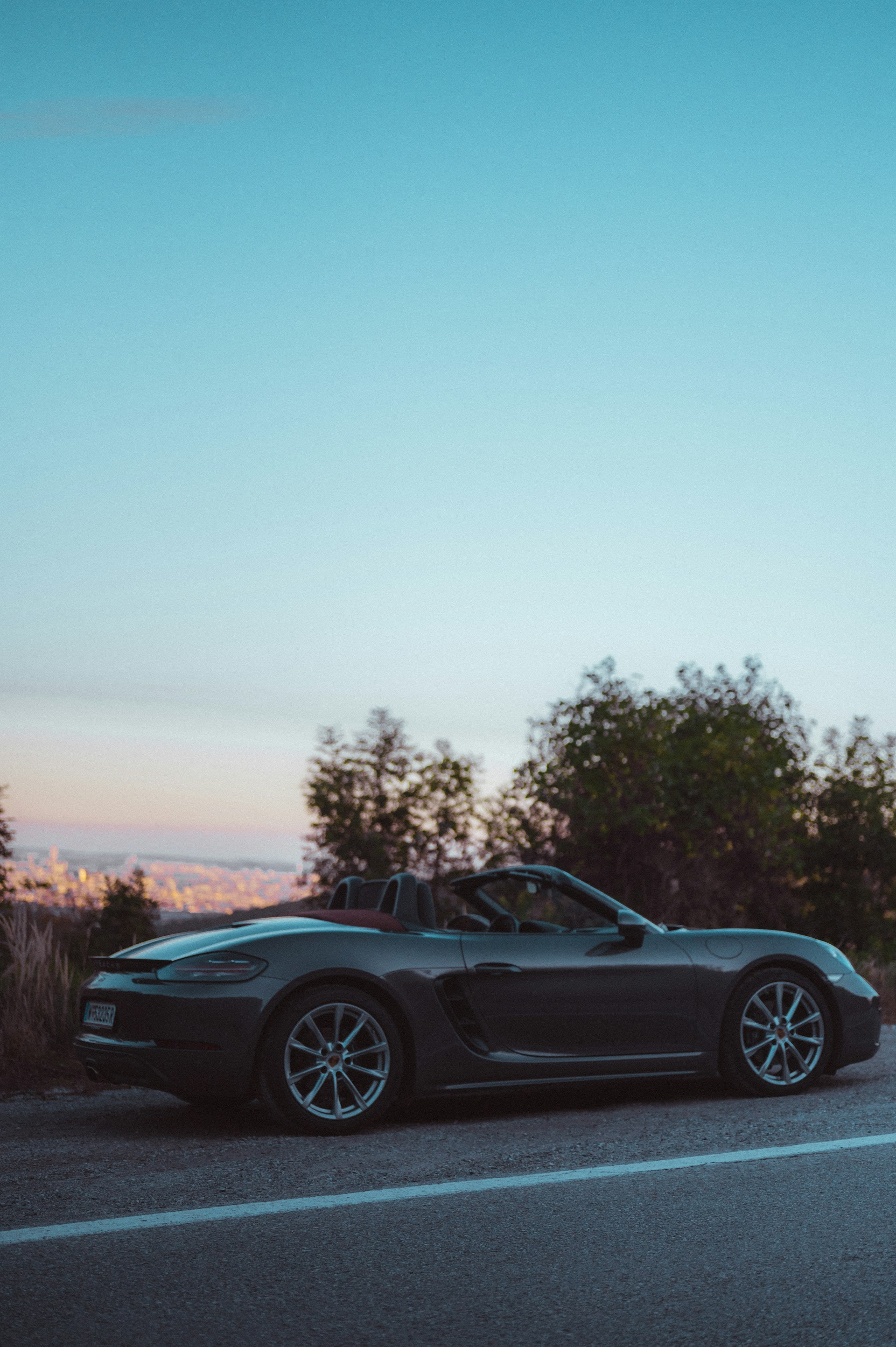 A luxury black convertible with the top down, parked in front of a modern city skyline at sunset.