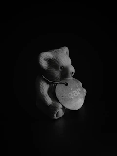 A small ceramic or plastic bear figurine is positioned against a dark background. The bear has a circular tag hanging around its neck with the word 'ear' printed on it.