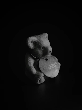 A small ceramic or plastic bear figurine is positioned against a dark background. The bear has a circular tag hanging around its neck with the word 'ear' printed on it.