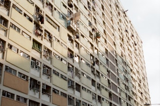A high-rise residential building with numerous windows and balconies, some featuring clothes hanging to dry. The architecture appears repetitive and the building stretches towards the sky at an angle, highlighting its height. The sunlight casts a soft glow on the surface, creating a mix of shadows and light.