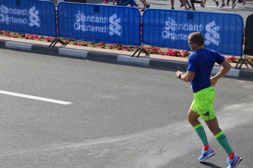 A man is running on a paved road near a barricaded section, possibly during a marathon or similar event. He wears bright neon green shorts, a blue shirt, and tall green compression socks. Nearby, blue barricades display the logo of Standard Chartered, and flowers line the pavement.