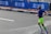 A man is running on a paved road near a barricaded section, possibly during a marathon or similar event. He wears bright neon green shorts, a blue shirt, and tall green compression socks. Nearby, blue barricades display the logo of Standard Chartered, and flowers line the pavement.