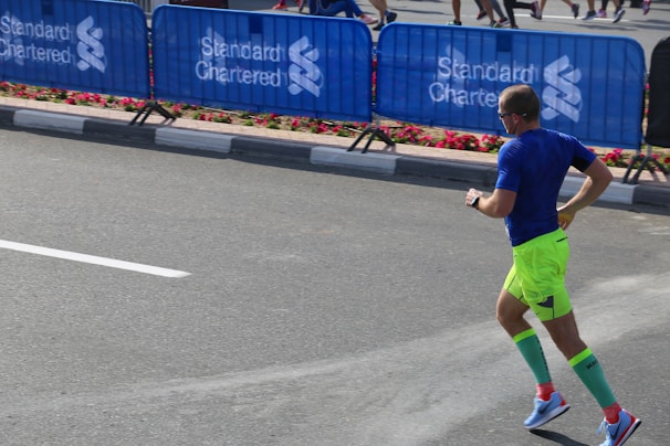 A man is running on a paved road near a barricaded section, possibly during a marathon or similar event. He wears bright neon green shorts, a blue shirt, and tall green compression socks. Nearby, blue barricades display the logo of Standard Chartered, and flowers line the pavement.