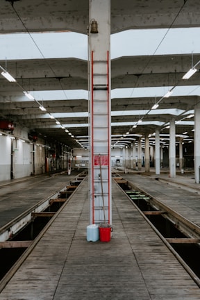 An industrial setting featuring a large, open space with evenly spaced concrete pillars and overhead fluorescent lighting. The floor is lined with wooden planks and a channel runs down the center, likely for vehicle maintenance. A ladder is centered on a pillar, with blue and red containers at its base.