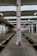 An industrial setting featuring a large, open space with evenly spaced concrete pillars and overhead fluorescent lighting. The floor is lined with wooden planks and a channel runs down the center, likely for vehicle maintenance. A ladder is centered on a pillar, with blue and red containers at its base.
