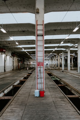An industrial setting featuring a large, open space with evenly spaced concrete pillars and overhead fluorescent lighting. The floor is lined with wooden planks and a channel runs down the center, likely for vehicle maintenance. A ladder is centered on a pillar, with blue and red containers at its base.