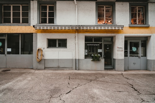 A building facade features a combination of textured white walls with a bold horizontal yellow stripe. Windows with gray frames are positioned symmetrically, and an awning with alternating light and dark stripes adds visual interest above the entrance. There is a neatly coiled hose hanging on the wall to the left, and a small potted plant is placed on the ground near the doorway.