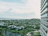 A view of a coastal resort area featuring multiple villas surrounded by lush greenery and palm trees. The image includes a body of water in the foreground and a large multi-story building to the right. In the background, the ocean extends to the horizon under a partly cloudy sky.