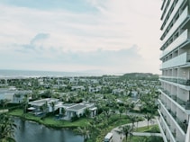 A view of a coastal resort area featuring multiple villas surrounded by lush greenery and palm trees. The image includes a body of water in the foreground and a large multi-story building to the right. In the background, the ocean extends to the horizon under a partly cloudy sky.