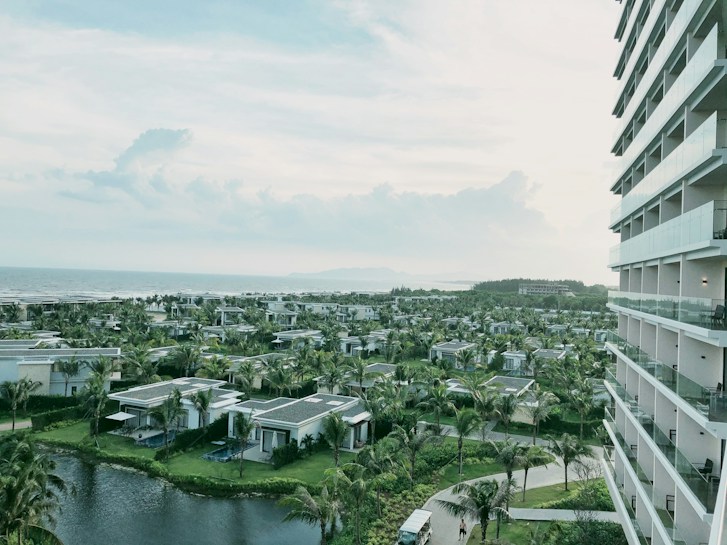 A view of a coastal resort area featuring multiple villas surrounded by lush greenery and palm trees. The image includes a body of water in the foreground and a large multi-story building to the right. In the background, the ocean extends to the horizon under a partly cloudy sky.