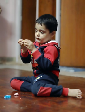 A young child sits on a wooden floor, dressed in a colorful outfit with red and blue patterns, intently examining an object in their hands. Around them, a few other small toys or objects are scattered on the ground. The background features a blurred doorway.
