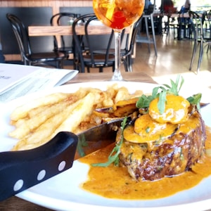 A plate of food featuring a steak covered in a rich orange sauce, garnished with arugula, and served with thick-cut fries. A sharp knife rests on the plate beside the steak. In the background, there is a blurred setting of a restaurant with wooden tables and black chairs. A tall glass containing an orange beverage with cherries is placed next to the plate.