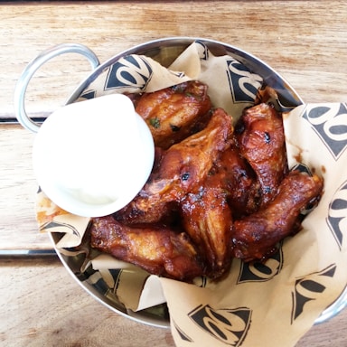 A metal bowl filled with crispy, golden-brown chicken wings arranged on a bed of brown paper with a decorative pattern. A small cup of creamy white sauce accompanies the wings, placed to the side. The setup is on a wooden table, enhancing the rustic and appetizing presentation.