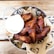 A metal bowl filled with crispy, golden-brown chicken wings arranged on a bed of brown paper with a decorative pattern. A small cup of creamy white sauce accompanies the wings, placed to the side. The setup is on a wooden table, enhancing the rustic and appetizing presentation.