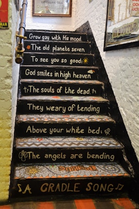 A set of stairs decorated with poetic phrases in white lettering against a black background. Each step features part of a poem with symbolic drawings like a sun, a figure of an angel, and other small icons. The walls around the stairs are adorned with posters and signs.