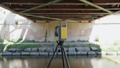 A yellow and black tripod-mounted surveying instrument is positioned under a metal bridge by a riverbank. The bridge has rust-colored girders and shadows cast across a concrete wall. Lush greenery grows along the banks with patches of sunlight highlighting parts of the scene.